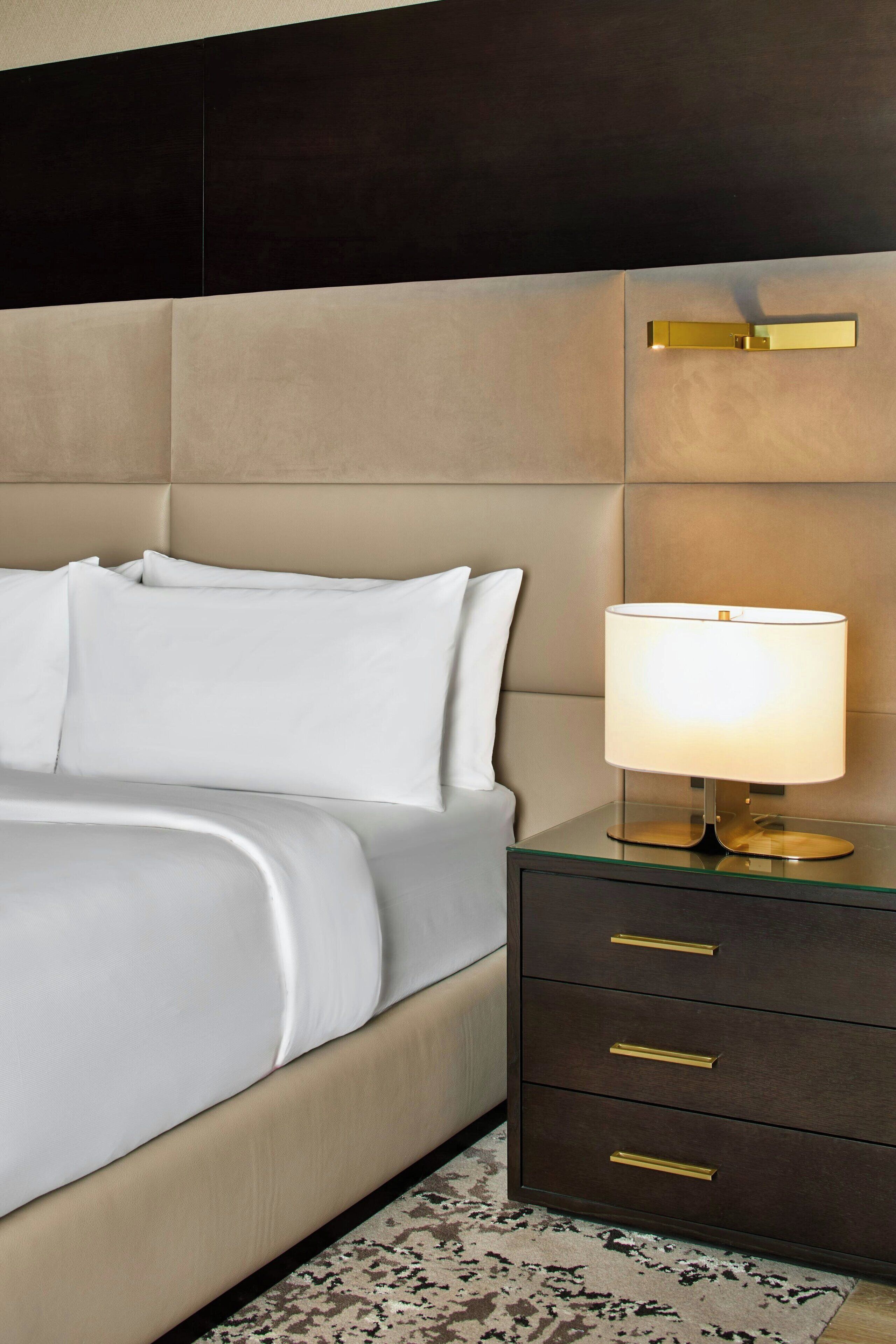 A neatly made bed with white linens, a bedside lamp with a gold base, and a dark brown nightstand with three drawers.