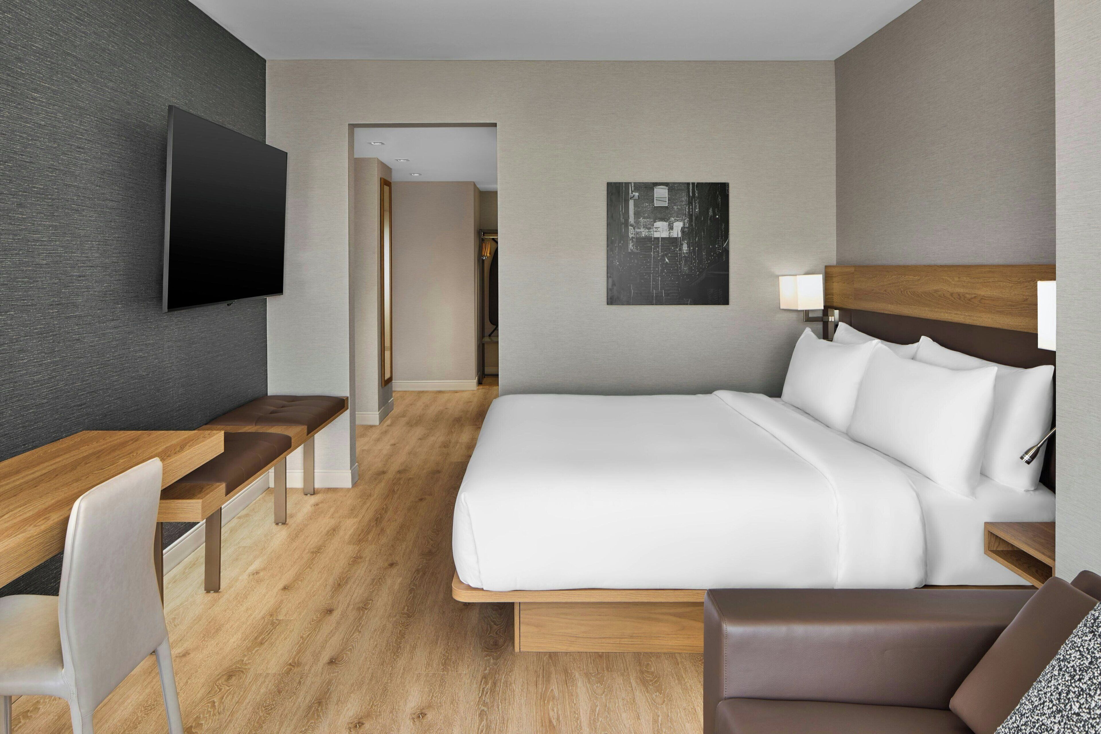 Una habitación de hotel moderna con una cama grande, un escritorio con una silla, un televisor de pantalla plana montado en la pared y un cuadro en la pared.