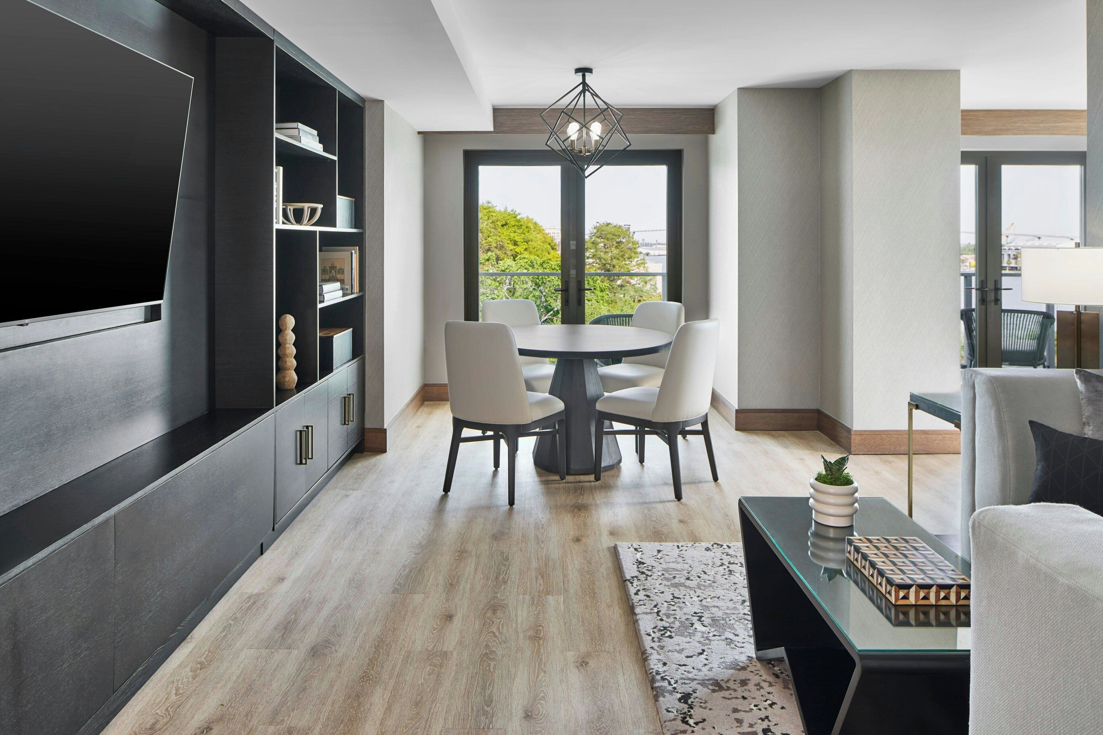 Una sala de estar moderna con zona de comedor, que presenta un televisor de pantalla plana grande, una mesa de comedor redonda con sillas, una mesa de centro de vidrio y un ventanal con vistas a zonas verdes.