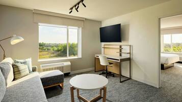 A modern hotel room with a large window, a flat-screen TV, a desk, a comfortable sofa, and a small round table.