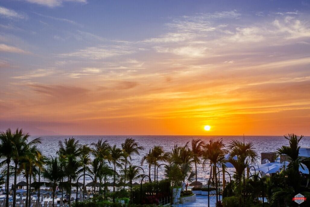Vista do pôr do sol sobre uma praia com palmeiras e guarda-sóis.
