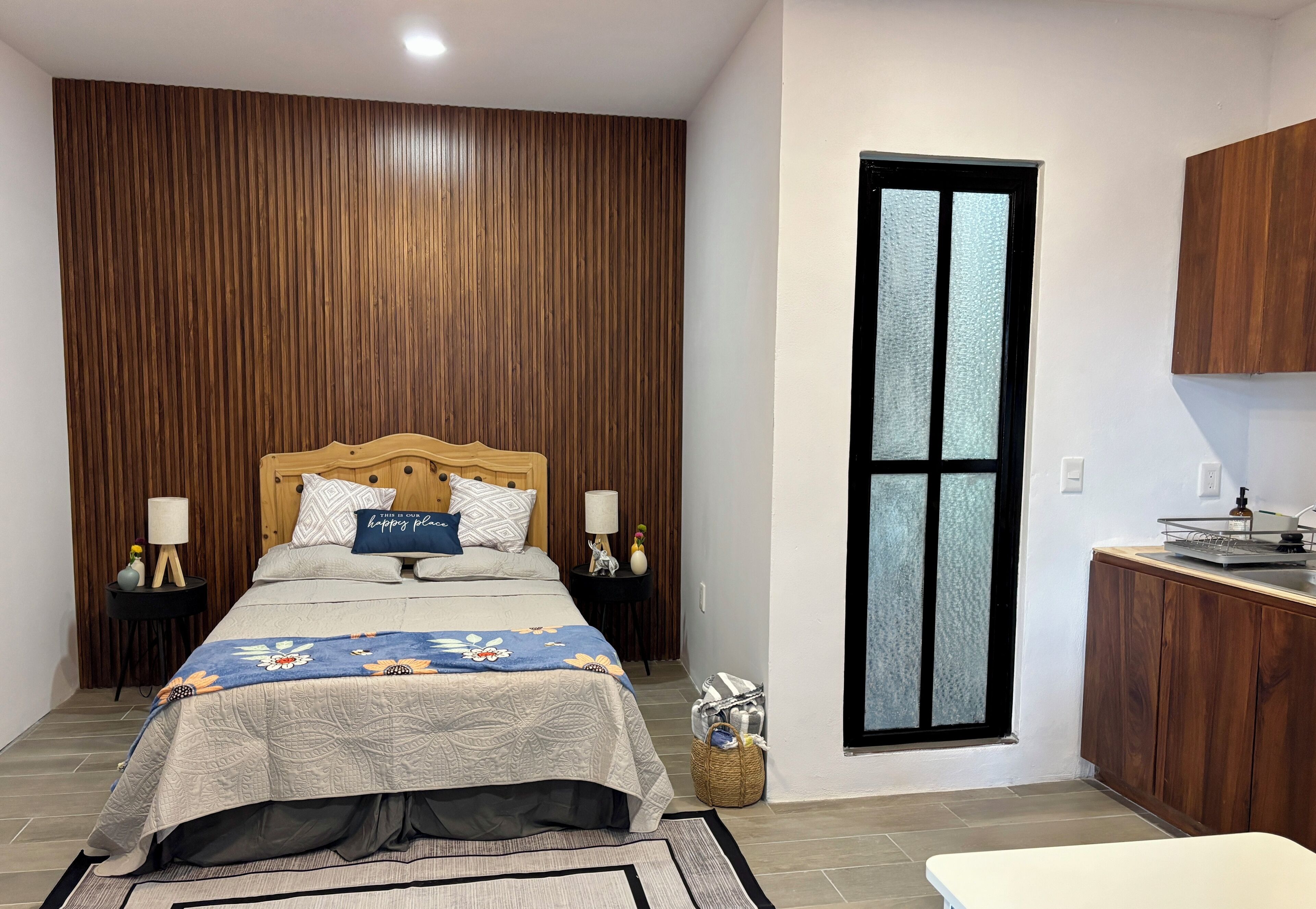 Quarto com cabeceira de madeira, cama grande com colcha floral, luminárias de cabeceira, mesa de cabeceira com abajur, pequena mesa com um vaso e janela com vidro fosco.