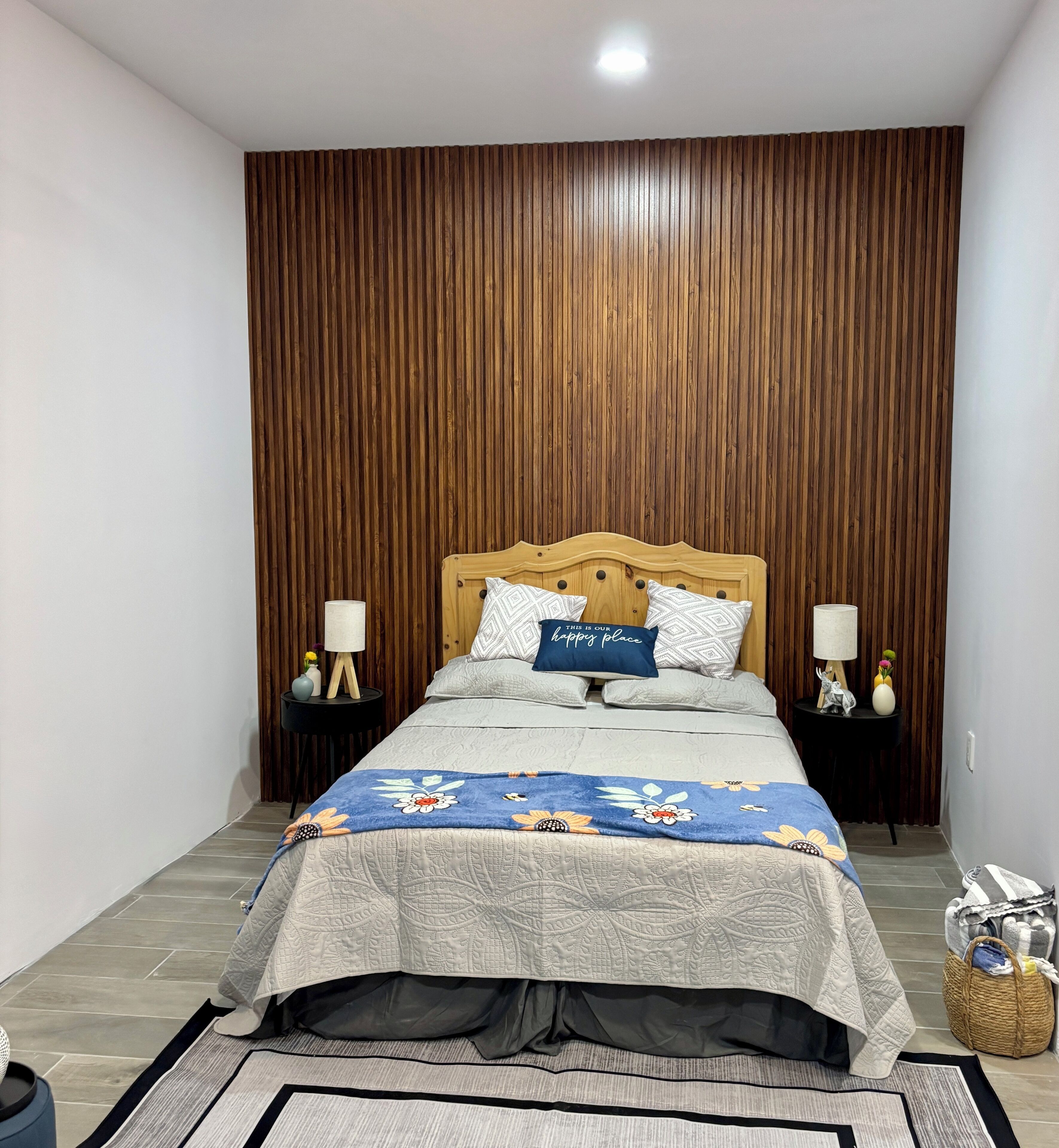 A bedroom with a wooden slat wall, a bed with a floral duvet, two bedside tables with lamps, and a small side table with decorative items.