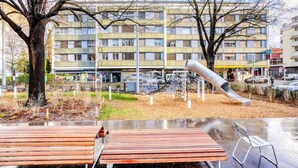 A playground with a slide and climbing structures, surrounded by benches and trees, in front of a multi-story building with shops and a car parked nearby. - Blueground | Gundeldingen 2bd apartment (Basel)