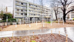 A playground with climbing structures and a fenced area, surrounded by a building with multiple windows and balconies. - Blueground | Gundeldingen 2bd apartment (Basel)