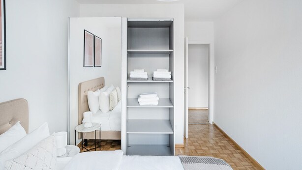 A bedroom with a bed, a bedside table, a chair, a wardrobe, and a hallway. - Blueground | Gundeldingen 2bd apartment (Basel)