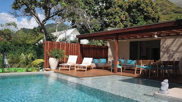 A swimming pool with lounge chairs and a covered seating area, surrounded by greenery and a mountain backdrop.