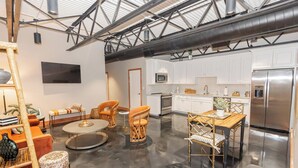 A spacious living area with a round coffee table, orange chairs, and a wooden dining table. A kitchen with white cabinets and stainless steel appliances is visible in the background.