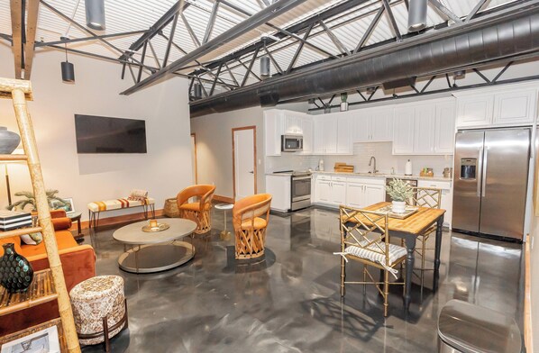 A spacious living area with a round coffee table, orange chairs, and a wooden dining table. A kitchen with white cabinets and stainless steel appliances is visible in the background.