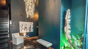 A modern lobby with a reflective floor, a white bench, a white armchair, a chandelier, a large abstract painting, and a green-lit wall with a plant.
