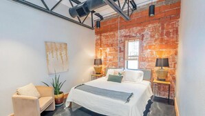 A modern bedroom with a brick wall, a bed with white bedding, two bedside tables with lamps, a sofa, and a potted plant.