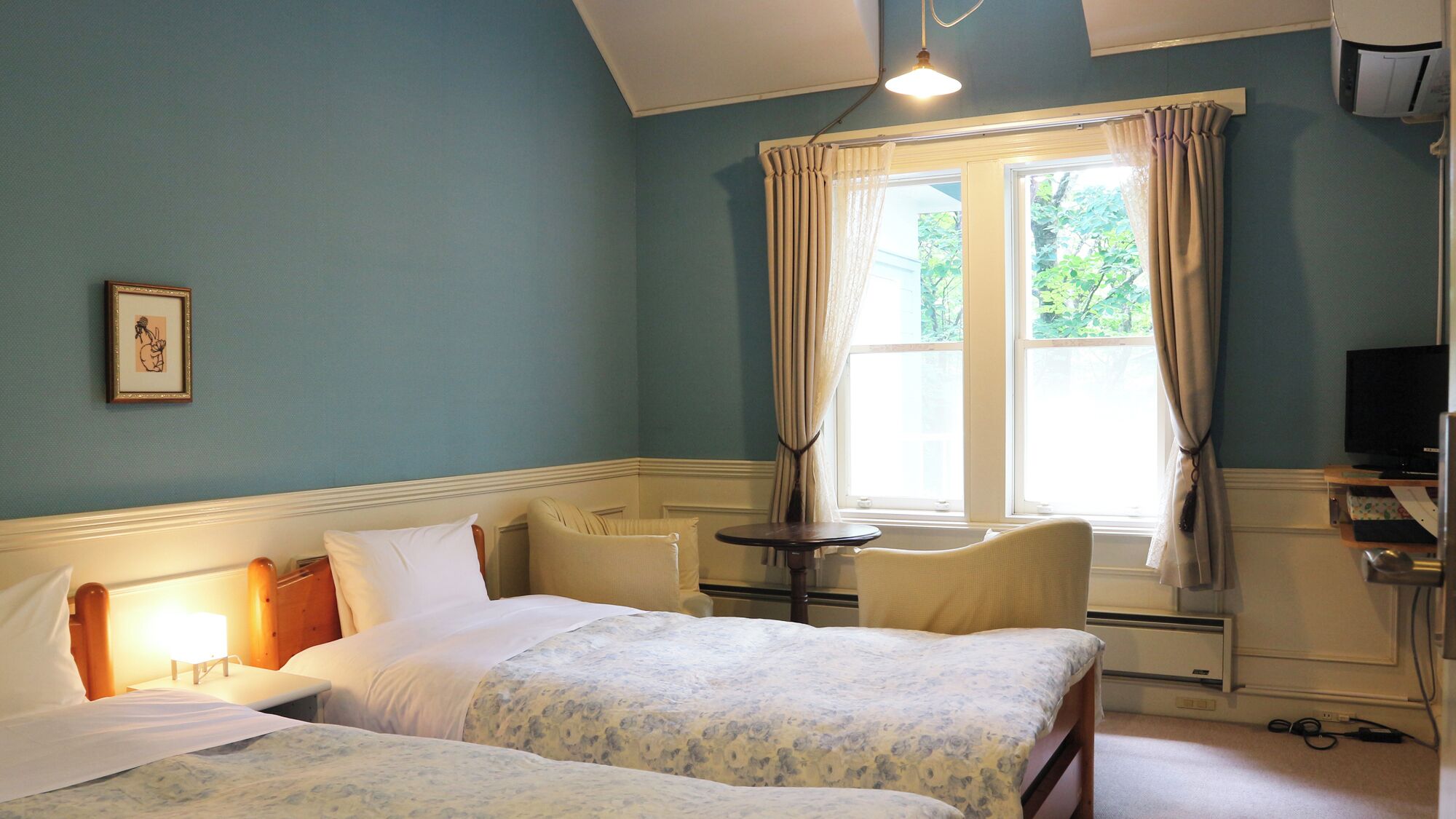 A hotel room with two beds, a small table, a TV, and a window with curtains.