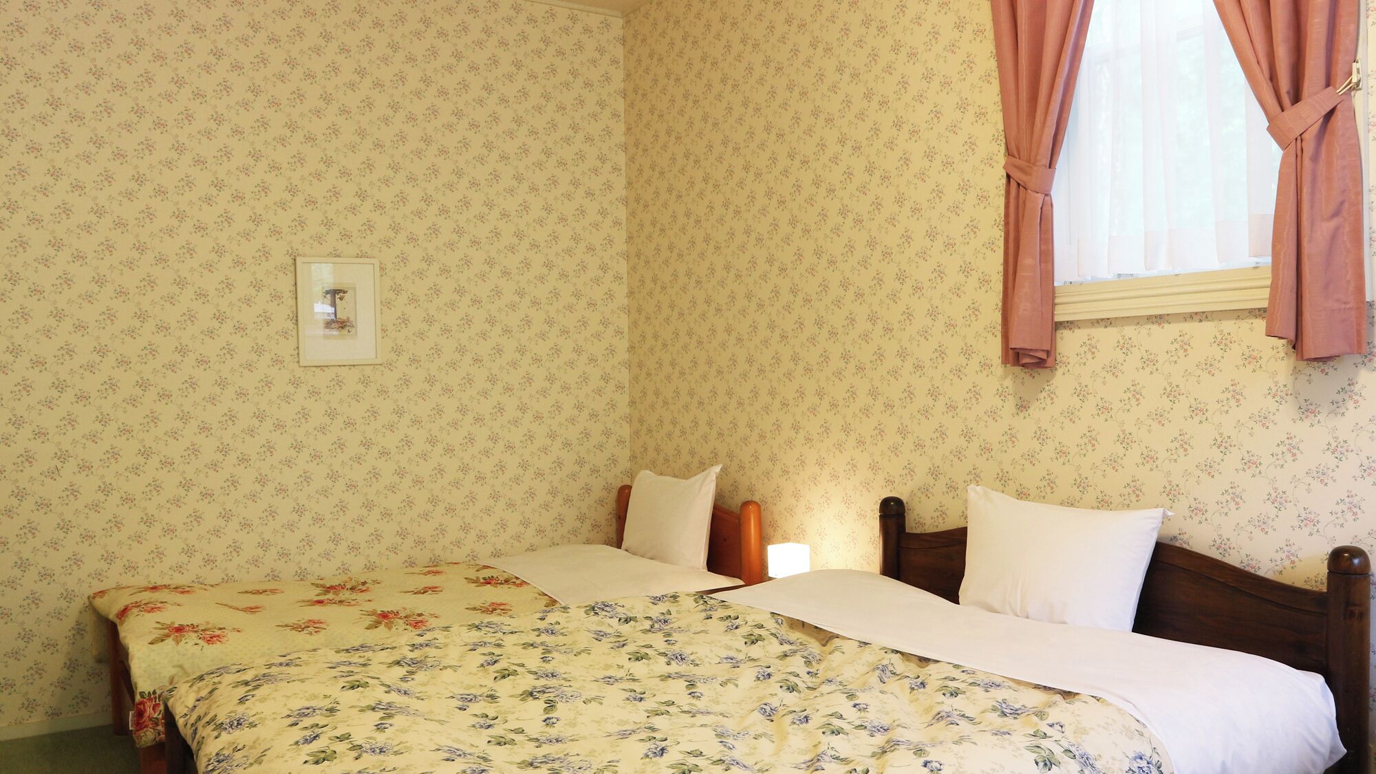 A bedroom with floral wallpaper, a bed with a floral bedspread, a nightstand, and a window with curtains.