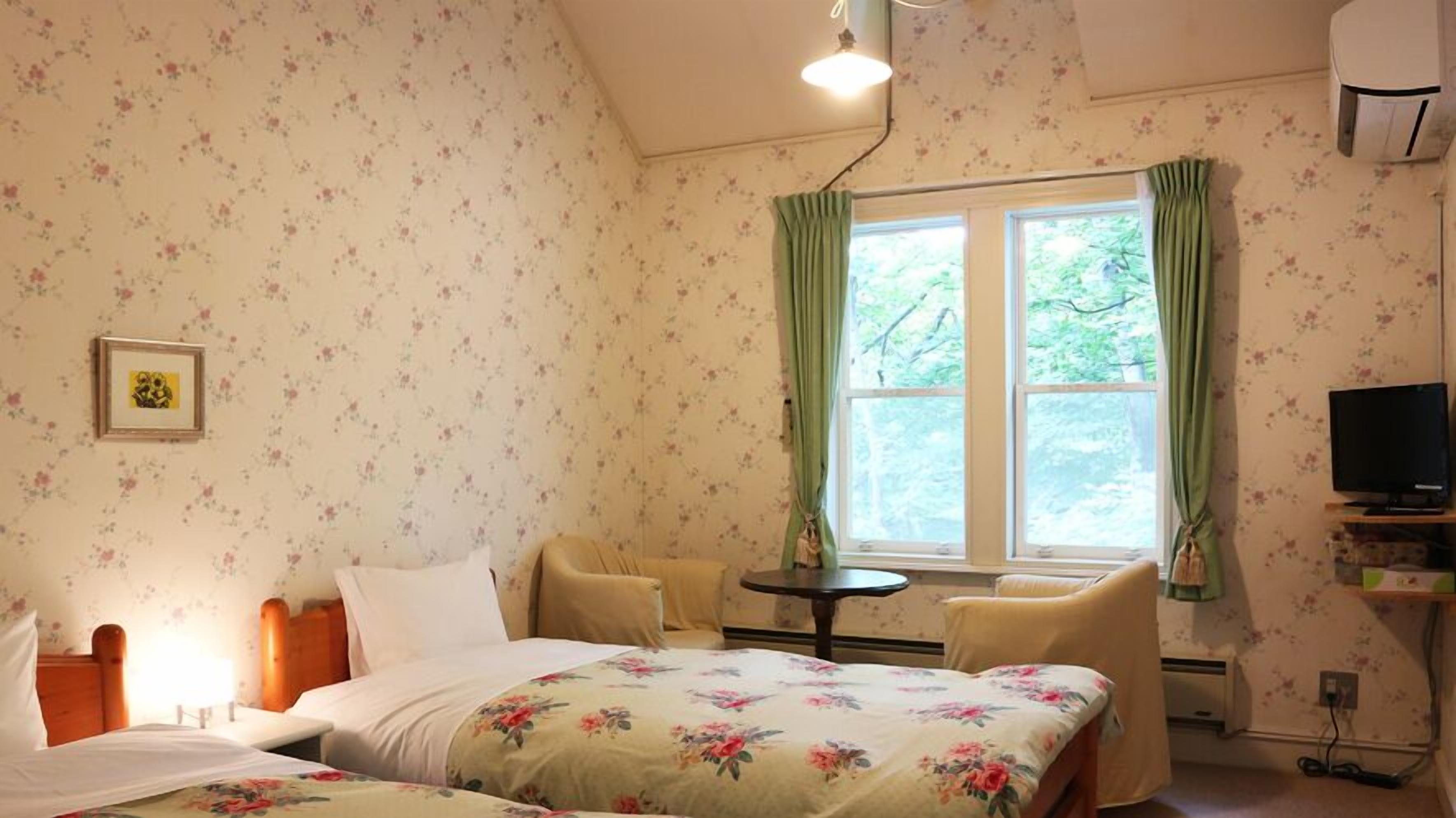 A bedroom with floral wallpaper, a bed with floral bedding, a chair, a small table, a window with curtains, and a television on a stand.
