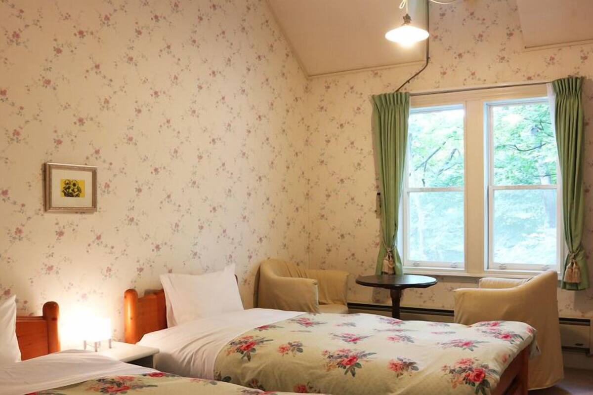A bedroom with floral wallpaper, a bed with floral bedding, a chair, a small table, a window with curtains, and a television on a stand.