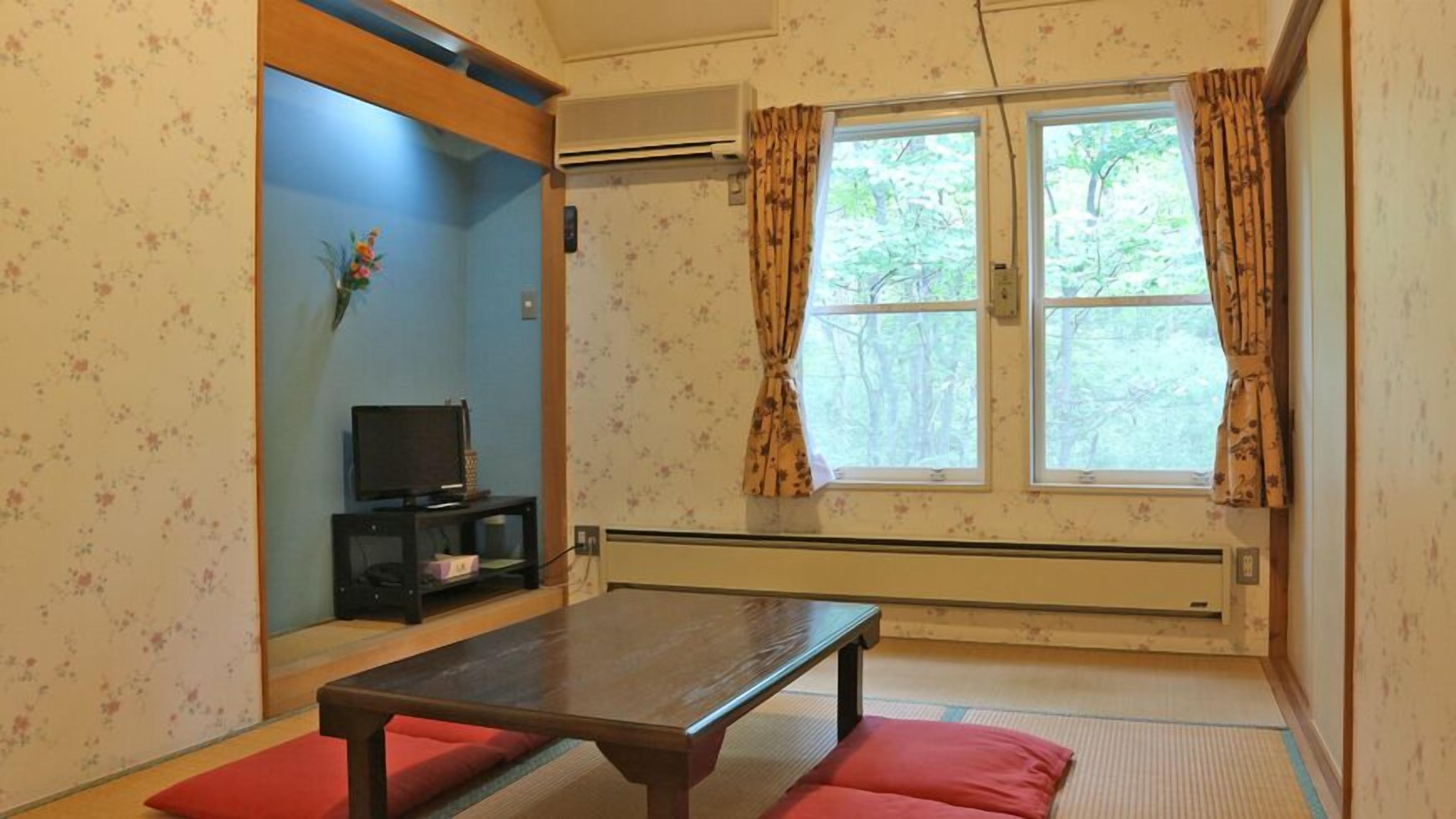 A room with a wooden table, red cushions, a TV on a stand, and a window with curtains.