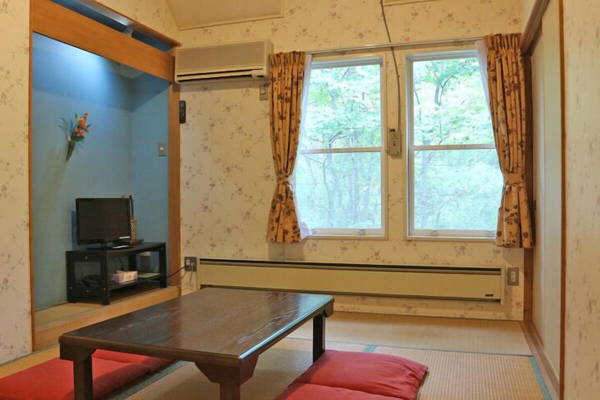 A room with a wooden table, red cushions, a TV on a stand, and a window with curtains.