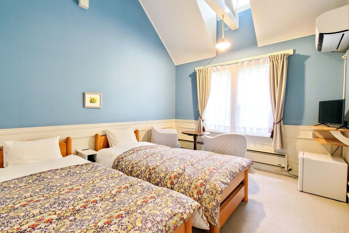 A bedroom with two beds, a desk, a chair, a TV, and a window with curtains.