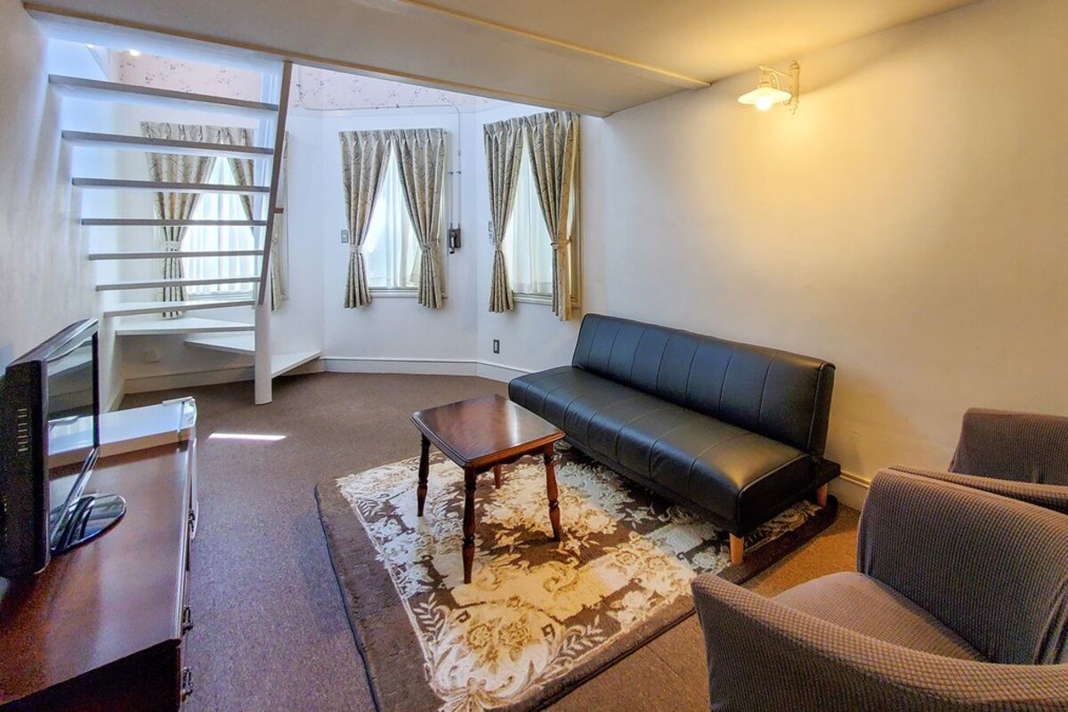A living room with a sofa, two armchairs, a coffee table, a TV, and a staircase leading to an upper level.