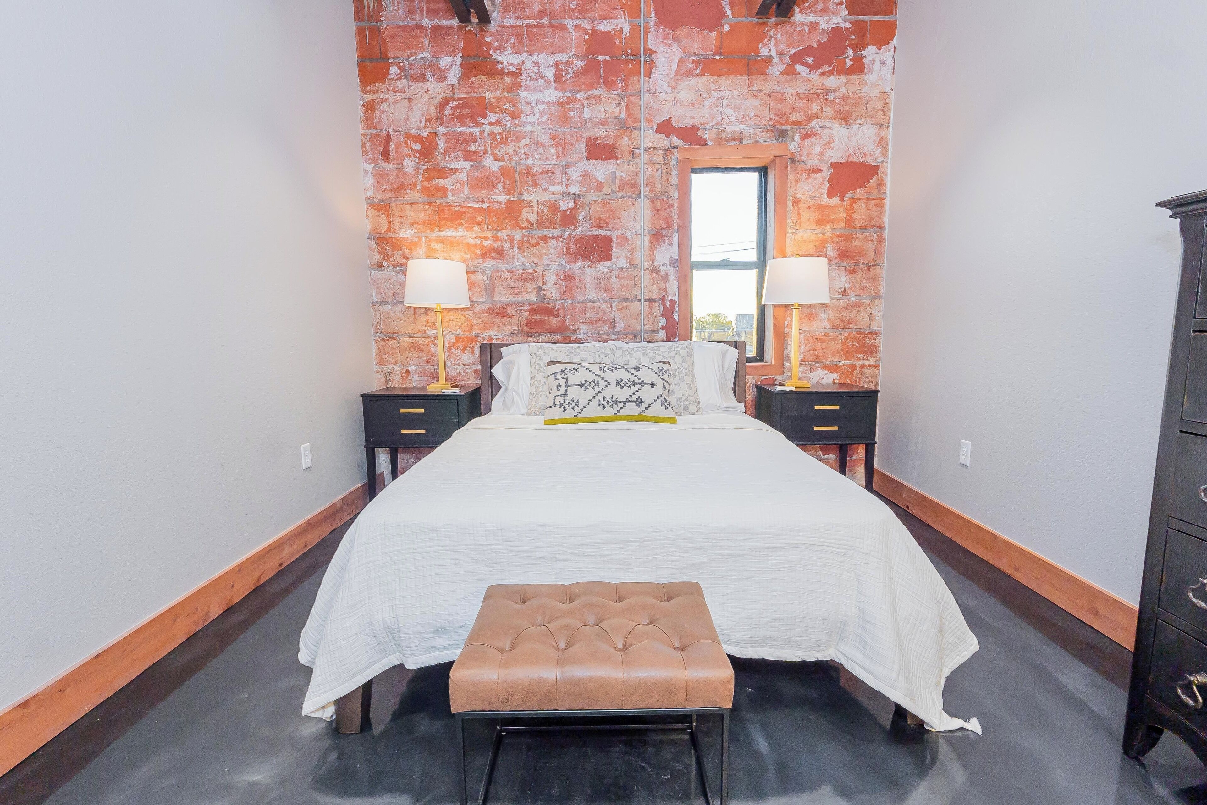 Une chambre avec un mur en briques, un lit recouvert d’une courtepointe blanche, deux tables de chevet avec des lampes, un banc en bois et une commode.
