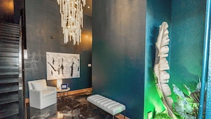 A modern lobby with a reflective floor, a white bench, a white armchair, a chandelier, a large abstract painting, and a green-lit wall with a plant.