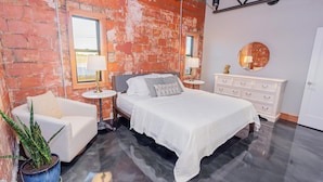 A bedroom with a brick wall, a bed with white bedding, a white dresser, and a round mirror on the wall.