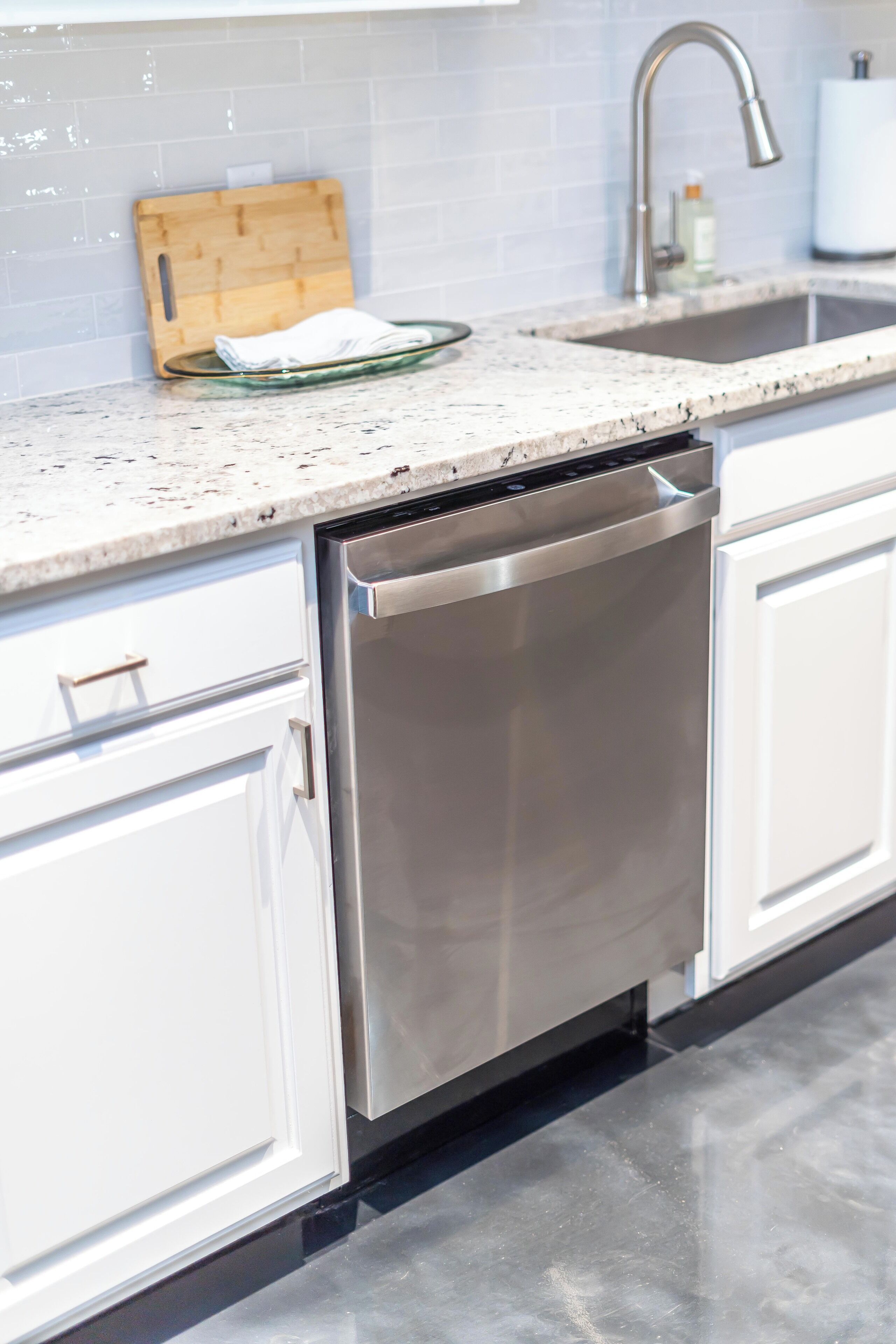 A modern kitchen with white cabinets, a stainless steel dishwasher, a granite countertop, and a wooden cutting board.
