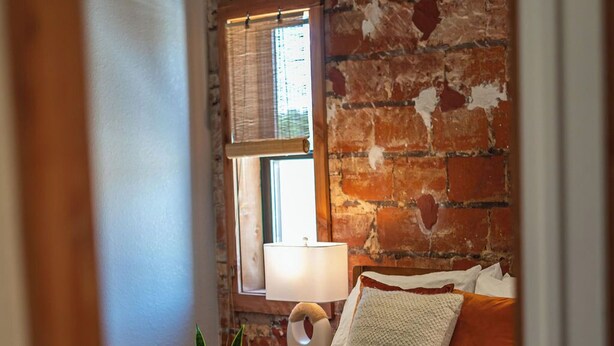A bedroom with a brick wall, a bed with white bedding, a bedside lamp, and a window with blinds.