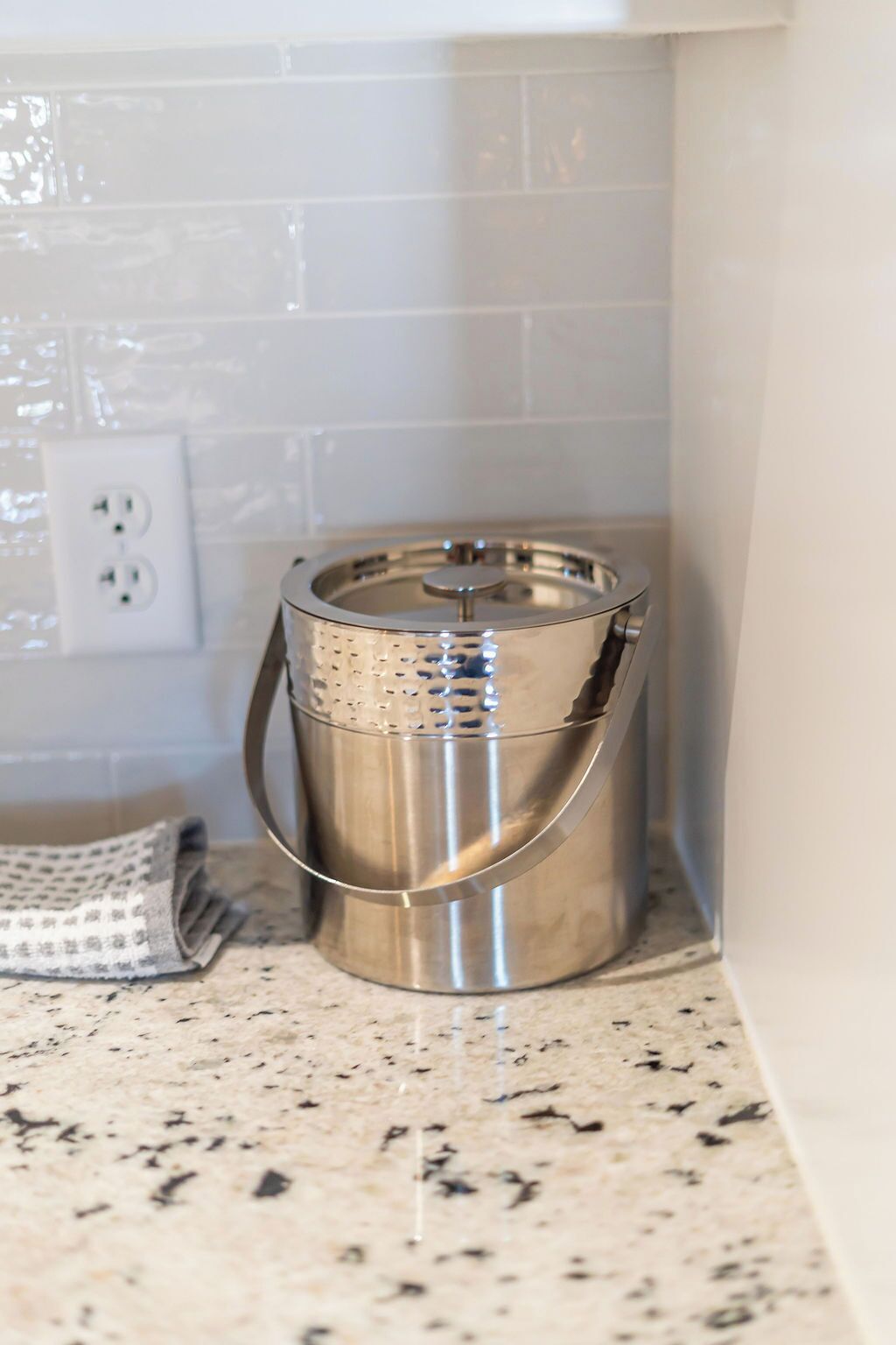 A stainless steel trash can with a perforated lid, placed on a speckled countertop near a white tiled wall and an electrical outlet.