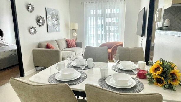 A dining area with a table set for four, surrounded by chairs. There is a sofa in the background, a television mounted on the wall, and a window with curtains.