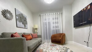 A living room with a grey sofa, a patterned rug, a brown armchair, a floor lamp, and a TV on the wall.