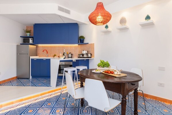 A compact kitchen with blue cabinets, a microwave, and a sink. There is a dining area with a table and chairs, and a decorative hanging light.