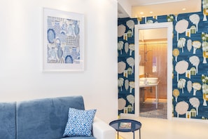 A modern living room with a blue sofa, a patterned pillow, and a small round table. A framed abstract artwork is on the wall, and a bathroom with a patterned wallpaper is visible through an open door.