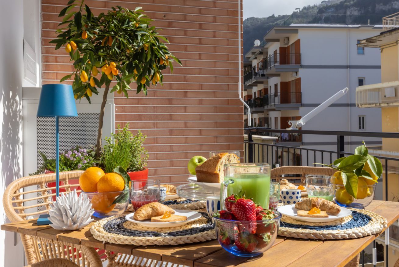 Un balcón con una mesa dispuesta para el desayuno, que incluye frutas, pan y una bebida.