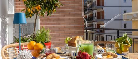 Un balcón con una mesa dispuesta para el desayuno, que incluye frutas, pan y una bebida.
