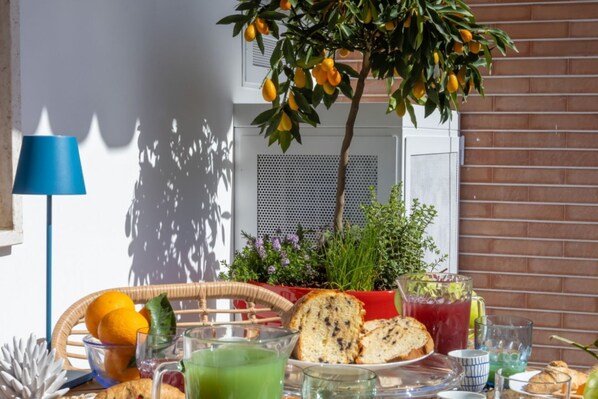 Desayuno con pan, fruta y bebidas sobre una mesa, con una planta en maceta y una lámpara al fondo.