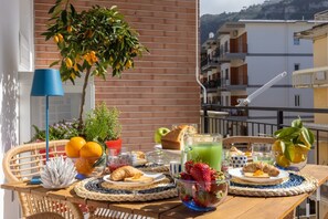 A balcony with a table set for breakfast, including fruits, bread, and a drink.