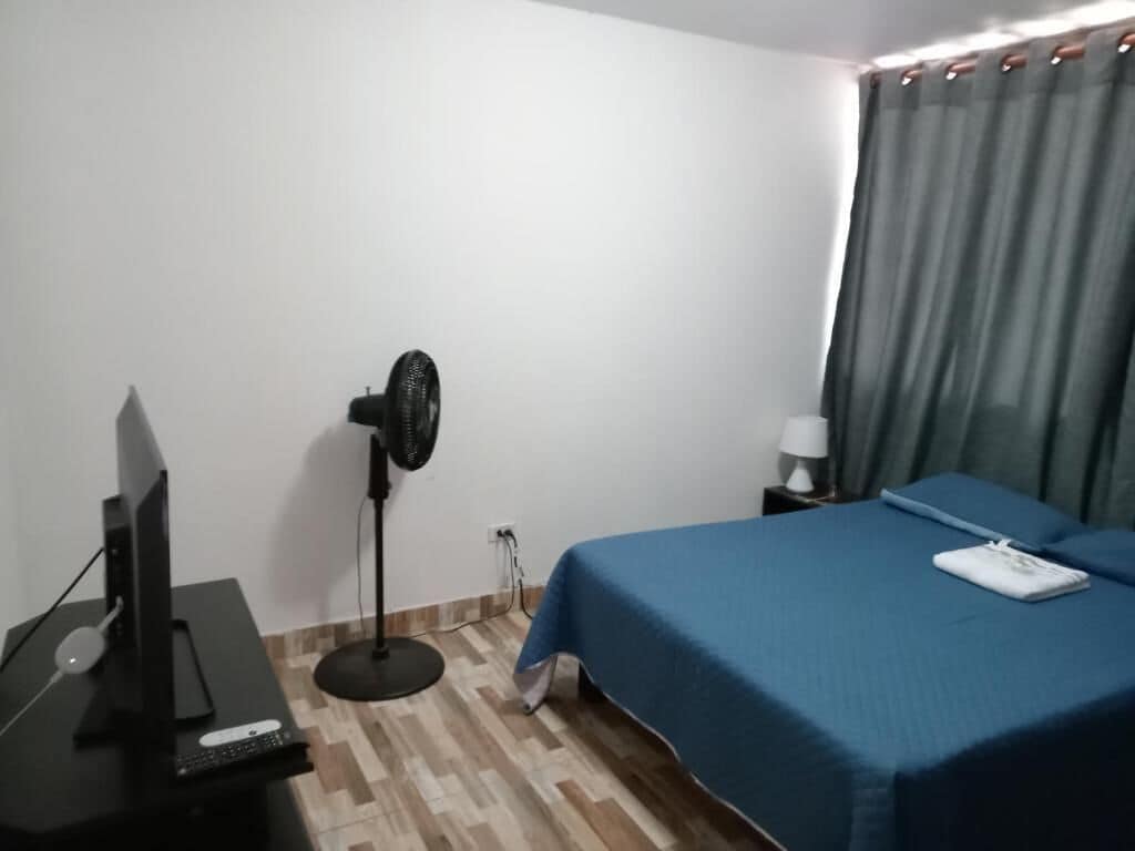 Una habitación de hotel con una cama, un escritorio con ordenador, un ventilador de pie y una mesita de noche con lámpara.