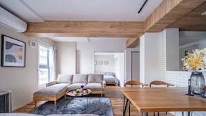 Een moderne woonkamer met een bank, poef en salontafel. Er staat een houten eettafel met stoelen. De kamer heeft houten balken in het plafond en een groot raam met gordijnen.
