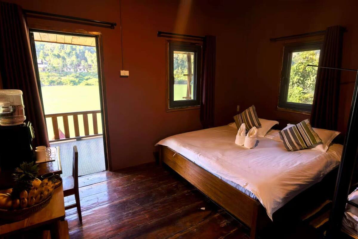 Ein Zimmer mit einem Bett, einem kleinen Tisch mit Obstschale, einem Stuhl und einem Fenster mit Blick auf Grünflächen.