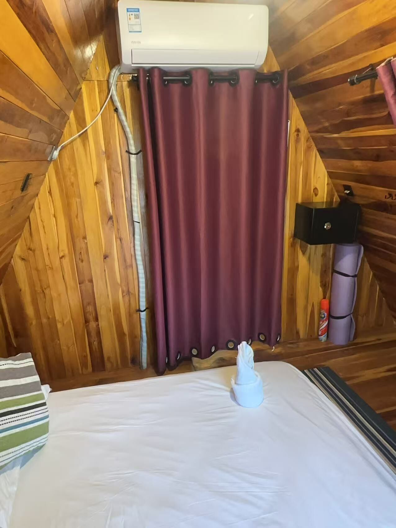 Ein Zimmer mit Holzwänden, einem Bett mit weißer Bettwäsche, einem roten Vorhang und einem weißen Klimagerät.