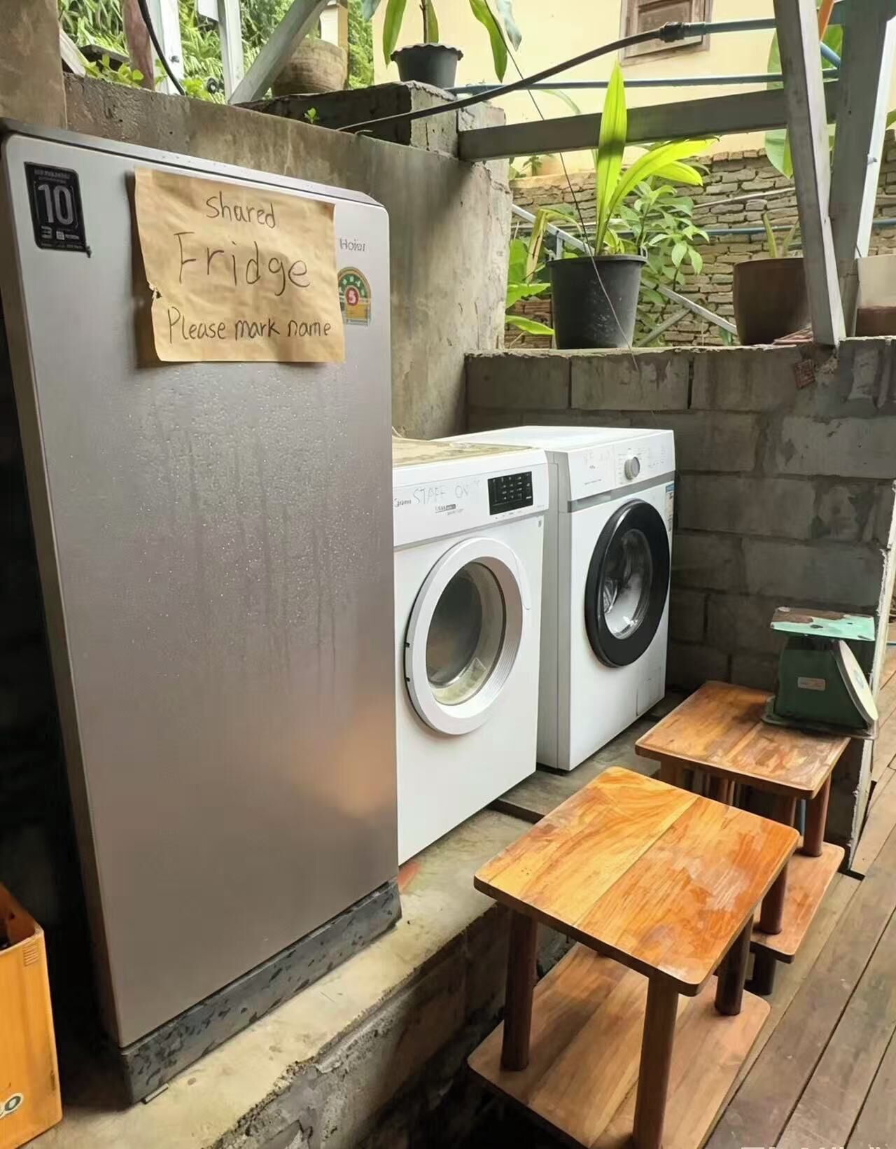 Un réfrigérateur à usage partagé avec un mot demandant aux utilisateurs d’indiquer ce qu’ils y ont mis, un lave-linge et des bancs en bois.