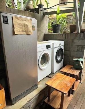 A shared refrigerator with a note requesting to mark it, a washing machine, and wooden benches.