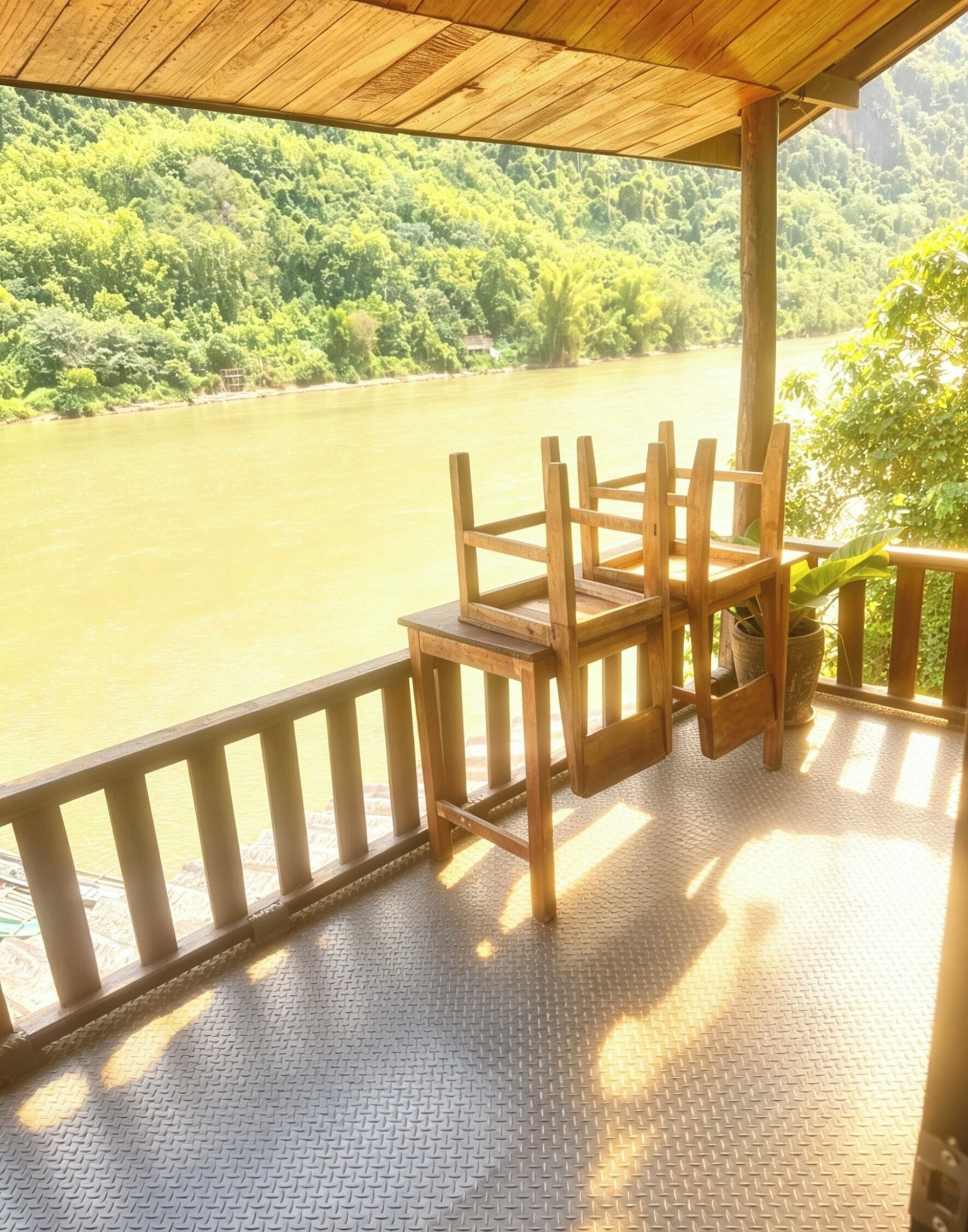 Une terrasse en bois avec une rambarde en métal, donnant sur une rivière, agrémentée de chaises et d’une plante en pot.
