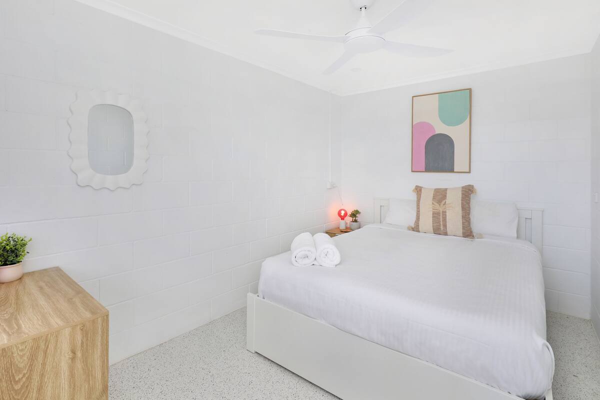 Un dormitorio blanco con cama, mesita de noche, planta en maceta, ventilador de techo y un cuadro colgado en la pared.