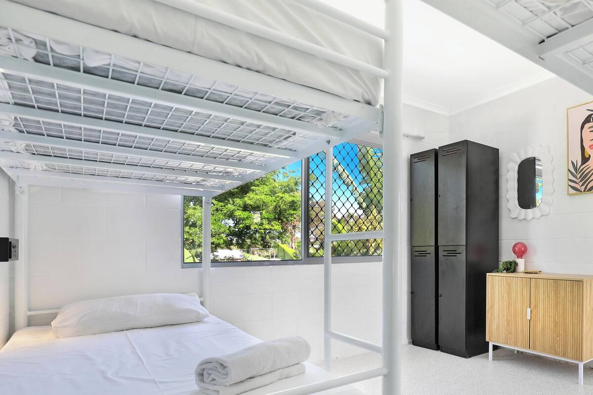 Una habitación con literas, un armario, un pequeño mueble de madera, un espejo y una ventana con vistas al verdor.