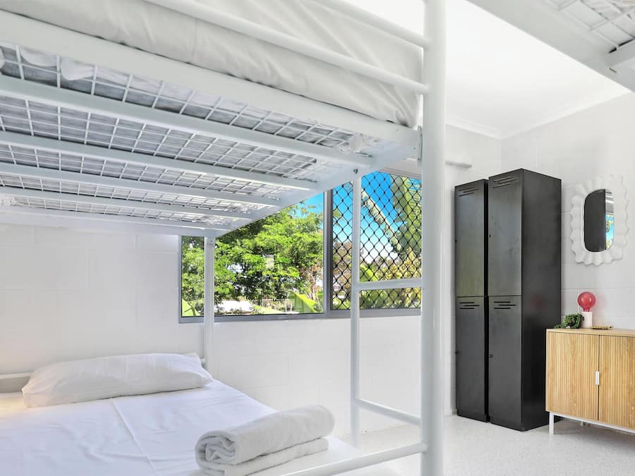Une chambre avec des lits superposés, une armoire, un petit meuble en bois, un miroir et une fenêtre donnant sur un paysage verdoyant.
