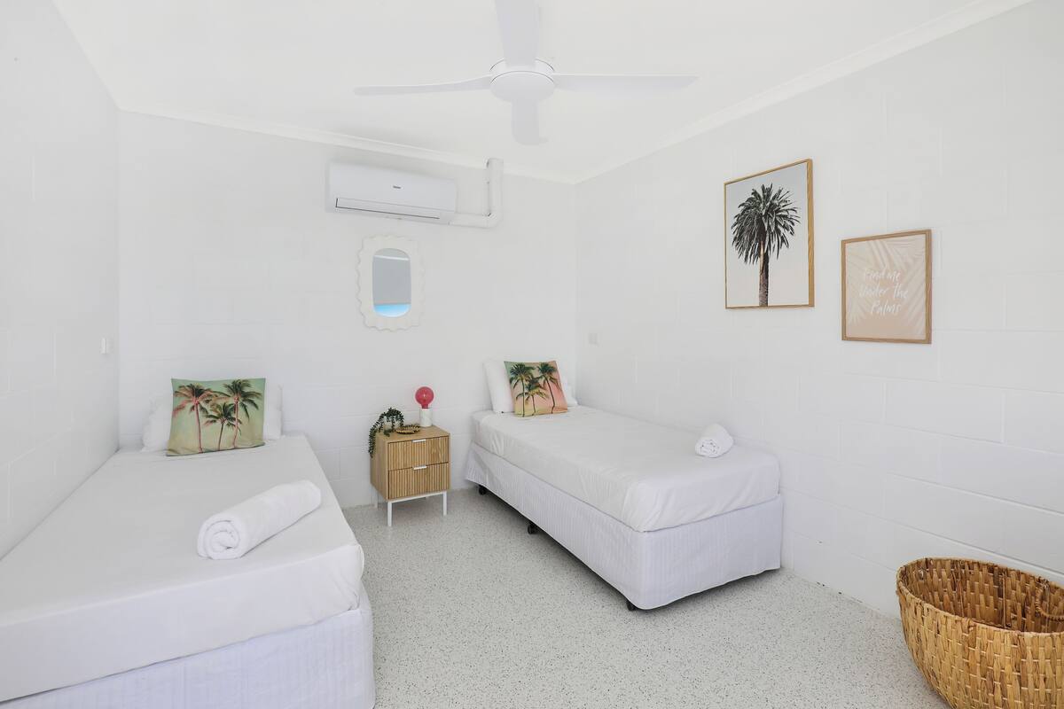 Una habitación con dos camas, ventilador de techo, unidad de aire acondicionado, mesita de noche de madera, espejo y una cesta tejida.