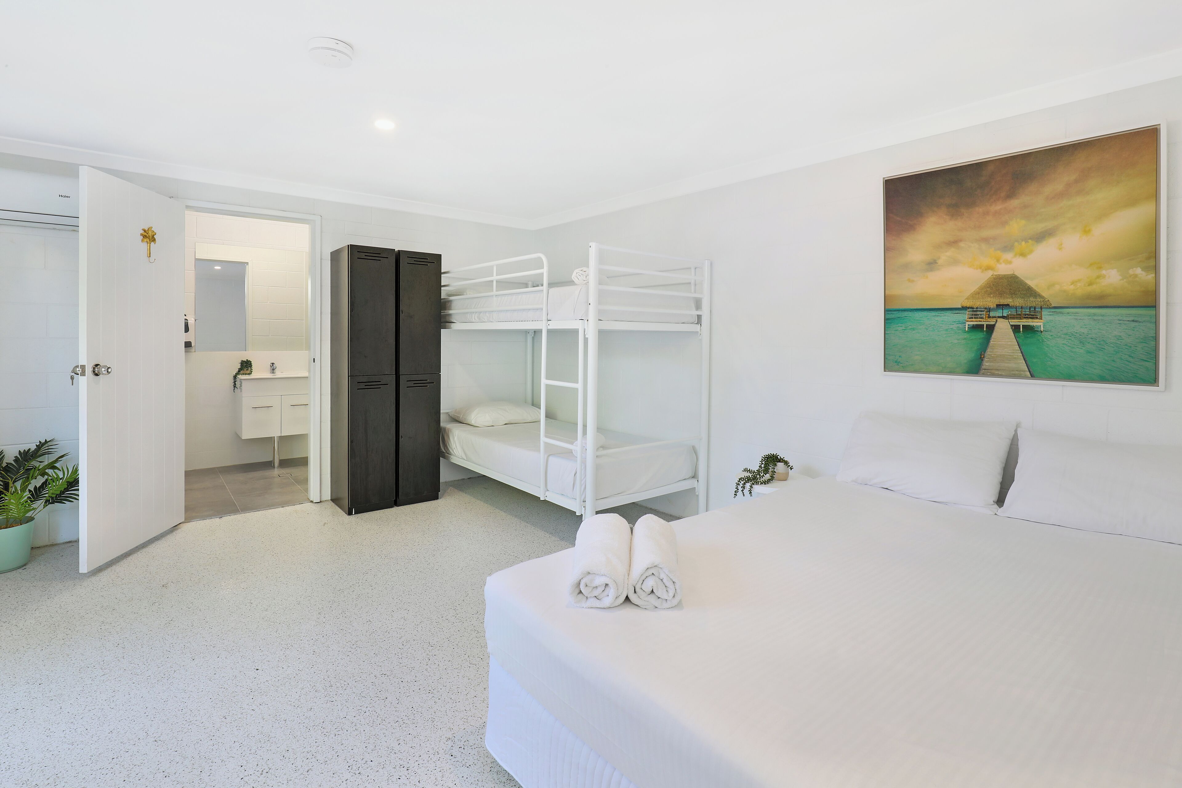Una camera d'albergo con un letto a castello, un letto singolo, un grande quadro raffigurante una scena tropicale, una pianta in vaso e un bagno con specchio e lavandino.