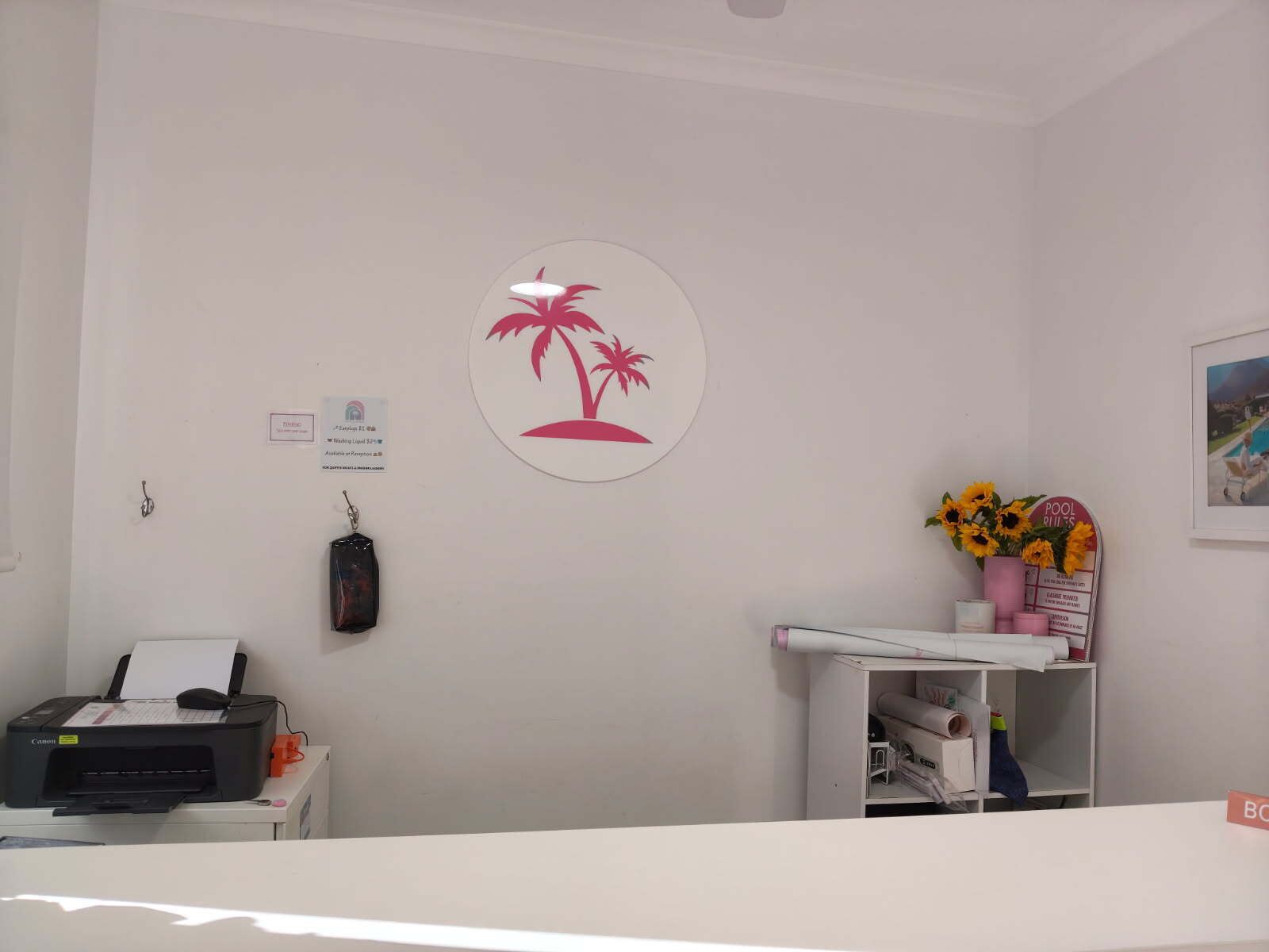 Zona de recepción con mostrador, una impresora, un cartel en la pared con palmeras y una estantería con girasoles.